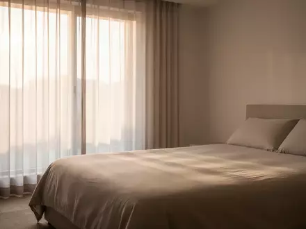 Ein minimalistisches Schlafzimmer mit sanftem Morgenlicht, das durch Vorhänge auf ein unberührtes Bett fällt. - Foto: über boerse-global.de
