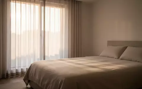 Ein minimalistisches Schlafzimmer mit sanftem Morgenlicht, das durch Vorhänge auf ein unberührtes Bett fällt. - Foto: über boerse-global.de