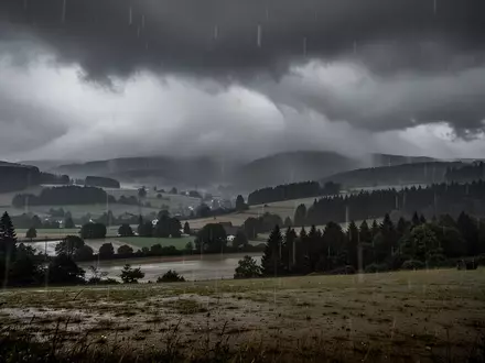 Eine stürmische, verregnete Landschaft bei düsterem Wetter mit tiefhängenden Wolken über einer hügeligen Waldregion. - Foto: über boerse-global.de