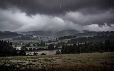 Eine stürmische, verregnete Landschaft bei düsterem Wetter mit tiefhängenden Wolken über einer hügeligen Waldregion. - Foto: über boerse-global.de