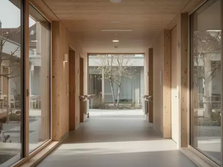 Ein ruhiger, von natürlichem Licht durchfluteter Flur in einem modernen Pflegeheim mit Blick auf einen begrünten Innenhof. - Foto: über boerse-global.de