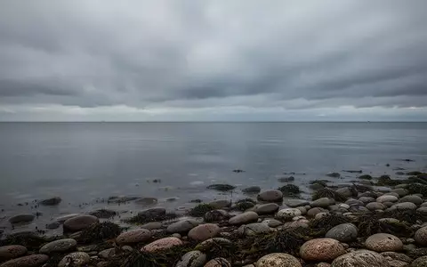 Die ruhige, graue Meeresoberfläche und die steinige Küste der Ostsee unter einem bewölkten Himmel. - Foto: über boerse-global.de