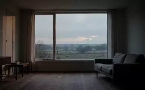 Ein minimalistisch eingerichtetes, leeres Wohnzimmer mit Blick auf eine ruhige, bewölkte Landschaft in der Dämmerung. - Foto: über boerse-global.de