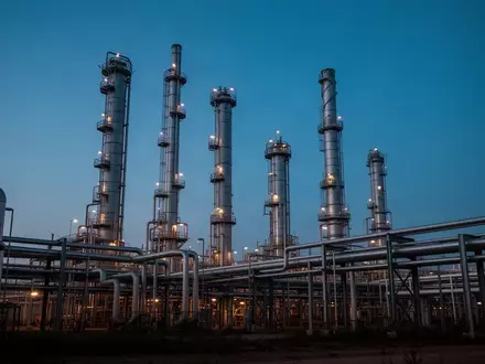 Eine industrielle Ölraffinerie bei Dämmerung mit hohen Destillationstürmen und Rohrleitungen vor einem blauen Himmel. - Foto: über boerse-global.de