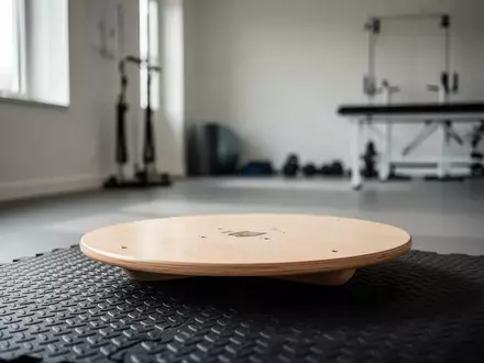 Ein hölzernes Balanceboard auf einer strukturierten Trainingsmatte in einem minimalistischen Physiotherapiestudio. - Foto: über boerse-global.de