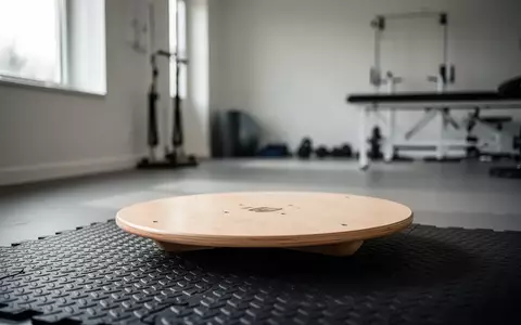 Ein hölzernes Balanceboard auf einer strukturierten Trainingsmatte in einem minimalistischen Physiotherapiestudio. - Foto: über boerse-global.de