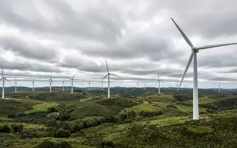 Moderne Windkraftanlagen in einer weitläufigen Landschaft bei bewölktem Himmel. - Foto: über boerse-global.de