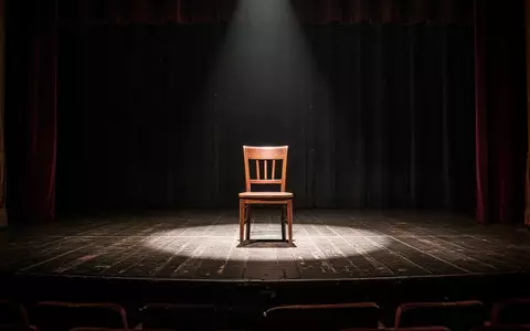 Eine leere Theaterbühne mit einem einzelnen Scheinwerfer und einem hölzernen Stuhl in einer nachdenklichen, schattigen Atmosphäre. - Foto: über boerse-global.de
