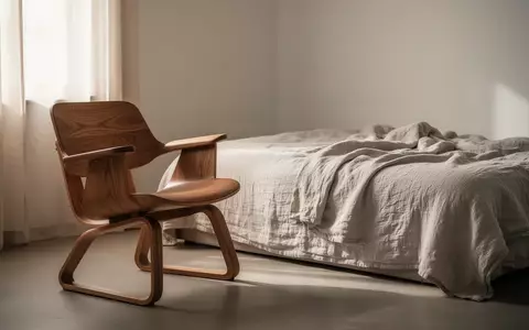 Ein minimalistisch eingerichteter, lichtdurchfluteter Raum mit einem ergonomischen Holzstuhl und einem ungemachten Bett. - Foto: über boerse-global.de