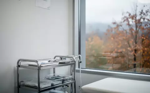Ein steriler, minimalistischer Untersuchungsraum in einer Klinik mit einem medizinischen Wagen vor einem Fenster mit Herbstlandschaft. - Foto: ĂĽber boerse-global.de