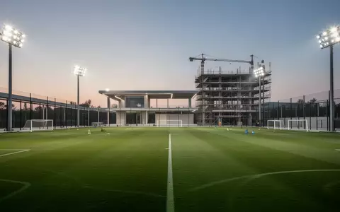 Ein modernes Fußball-Trainingsgelände mit einem grünen Spielfeld und einem im Bau befindlichen Gebäude unter Flutlicht. - Foto: über boerse-global.de