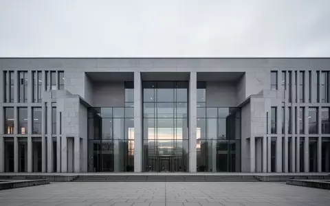 Moderne, minimalistische Architektur eines öffentlichen Gebäudes mit grauer Steinfassade und Glasfront unter bewölktem Himmel. - Foto: über boerse-global.de