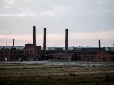 Eine weitläufige, menschenleere Industrielandschaft mit alten Fabrikgebäuden und Schornsteinen vor einem bewölkten Himmel. - Foto: über boerse-global.de