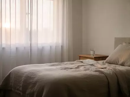 Ein minimalistisch eingerichtetes Schlafzimmer in der Morgendämmerung mit weichem Lichteinfall durch helle Vorhänge. - Foto: über boerse-global.de