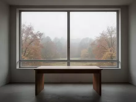 Ein minimalistischer, leerer Raum mit einem hölzernen Schreibtisch vor einem Fenster, das auf eine herbstliche Landschaft blickt. - Foto: über boerse-global.de