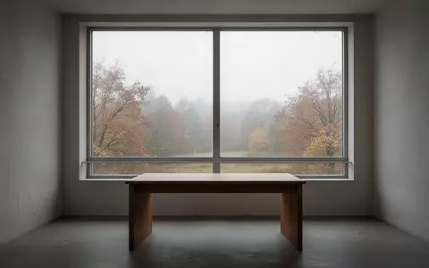 Ein minimalistischer, leerer Raum mit einem hölzernen Schreibtisch vor einem Fenster, das auf eine herbstliche Landschaft blickt. - Foto: über boerse-global.de