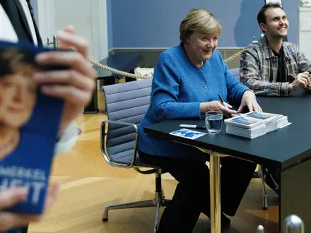 Merkel signiert Kunstpostkarten im Bode-Museum neben dem Maler ihres dort ausgestellten Porträts, Jérémie Queyras.  - Foto: Carsten Koall/dpa