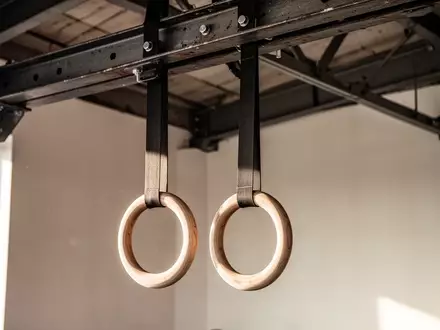 Zwei hölzerne Gymnastikringe hängen an stabilen Gurten von einer Industriedecke in einem minimalistischen Trainingsraum. - Foto: über boerse-global.de
