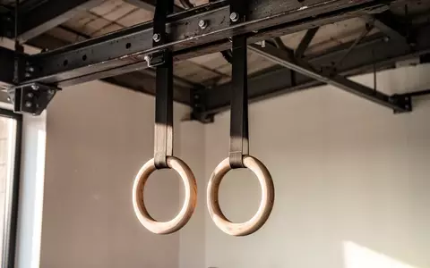 Zwei hölzerne Gymnastikringe hängen an stabilen Gurten von einer Industriedecke in einem minimalistischen Trainingsraum. - Foto: über boerse-global.de