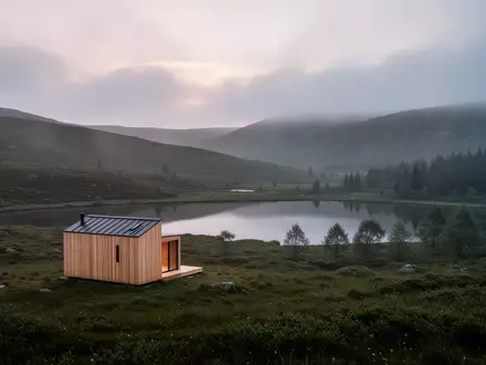 Eine einsame Holzhütte in einer nebligen, schottischen Hügellandschaft mit einem stillen See bei Dämmerung. - Foto: über boerse-global.de