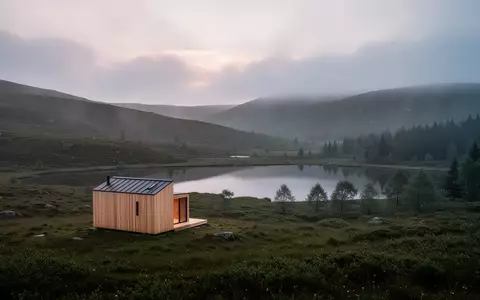 Eine einsame Holzhütte in einer nebligen, schottischen Hügellandschaft mit einem stillen See bei Dämmerung. - Foto: über boerse-global.de