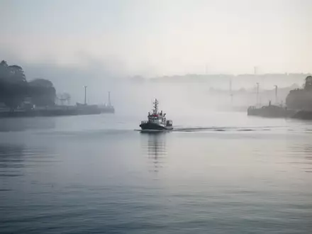Ein ruhiges Hafenbecken im morgendlichen Nebel mit einem in der Ferne fahrenden Lotsenboot. - Foto: über boerse-global.de