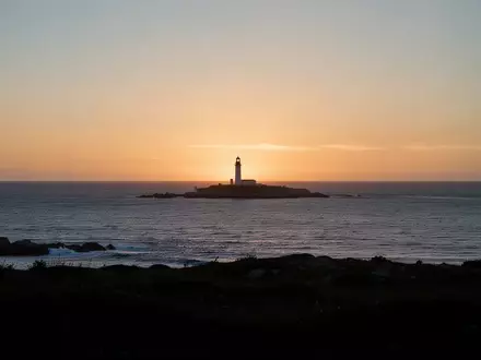 Eine ruhige Küstenlandschaft mit einem fernen Leuchtturm bei Sonnenuntergang über dem Meer. - Foto: über boerse-global.de
