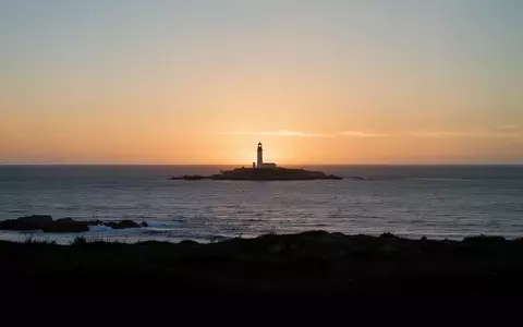 Eine ruhige Küstenlandschaft mit einem fernen Leuchtturm bei Sonnenuntergang über dem Meer. - Foto: über boerse-global.de