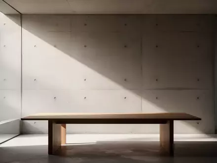 Ein minimalistischer, lichtdurchfluteter Raum mit einer schlichten Betonwand und einem leeren Holztisch in einer ruhigen Atmosphäre. - Foto: über boerse-global.de
