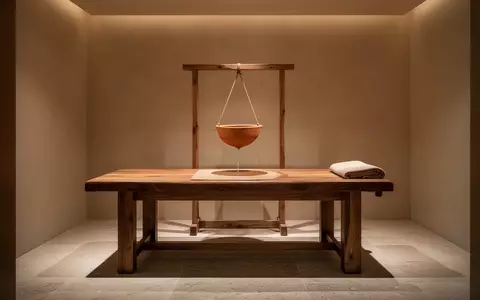 Ein ruhiger, minimalistischer Behandlungsraum mit einer hölzernen Massageliege und einer Vorrichtung für eine Shirodhara-Therapie. - Foto: über boerse-global.de