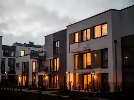 Moderne Wohngebäude in der Abenddämmerung mit warm beleuchteten Fenstern vor einem bewölkten Himmel. - Foto: über boerse-global.de
