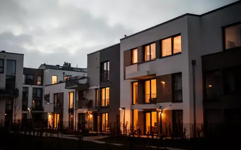 Moderne Wohngebäude in der Abenddämmerung mit warm beleuchteten Fenstern vor einem bewölkten Himmel. - Foto: über boerse-global.de