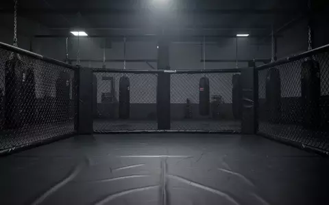 Ein leerer MMA-Trainingsring mit schwarzem Boden und Metallzaun in einem minimalistischen, abgedunkelten Fitnessstudio. - Foto: über boerse-global.de