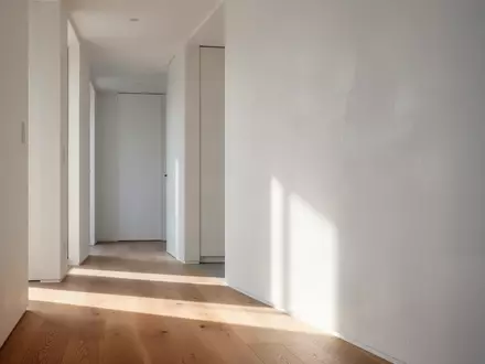 Ein minimalistischer, moderner Wohnungsflur mit klaren weißen Wänden und natürlichem Licht, das sanfte Schatten auf einen Eichenholzboden wirft. - Foto: über boerse-global.de