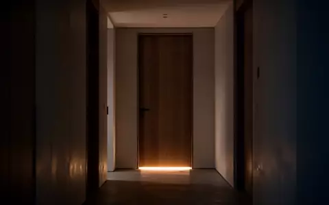 Ein schmaler Flur bei Nacht mit einer geschlossenen Zimmertür, unter der ein sanfter Lichtschein hervordringt. - Foto: über boerse-global.de