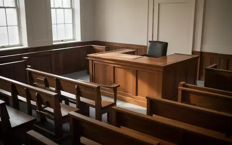 Ein leerer, sonnendurchfluteter Gerichtssaal mit hölzernen Sitzbänken und Richtertisch. - Foto: über boerse-global.de