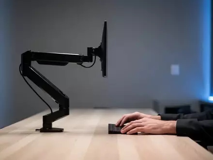 Ein minimalistischer, moderner Arbeitsplatz mit einem schwarzen Monitorarm an einem hellen Holztisch vor einer dezent beleuchteten Wand. - Foto: über boerse-global.de