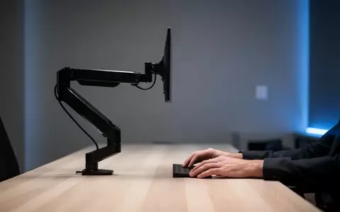 Ein minimalistischer, moderner Arbeitsplatz mit einem schwarzen Monitorarm an einem hellen Holztisch vor einer dezent beleuchteten Wand. - Foto: ĂĽber boerse-global.de