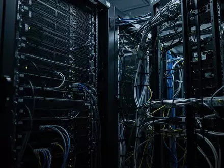 Dunkler Serverraum mit metallischen Rack-Schränken und komplexer Kabelführung in einem industriellen Umfeld. - Foto: über boerse-global.de