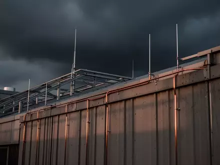 Metallische Blitzschutzvorrichtungen und Erdungsleitungen auf dem Dach eines modernen Industriegebäudes bei bewölktem Himmel. - Foto: über boerse-global.de
