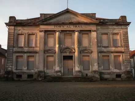 Ein verlassenes und sanierungsbedürftiges Rathausgebäude auf einem leeren Stadtplatz bei Dämmerung. - Foto: über boerse-global.de