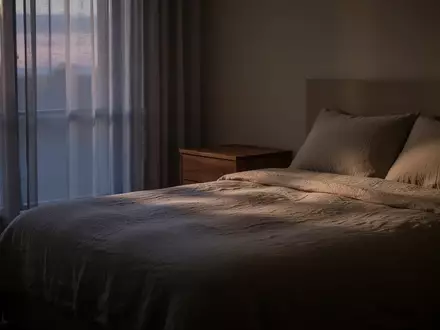 Ein minimalistisch eingerichtetes, ruhiges Schlafzimmer in der Abenddämmerung mit weichem Licht und einer ordentlichen Bettdecke. - Foto: über boerse-global.de