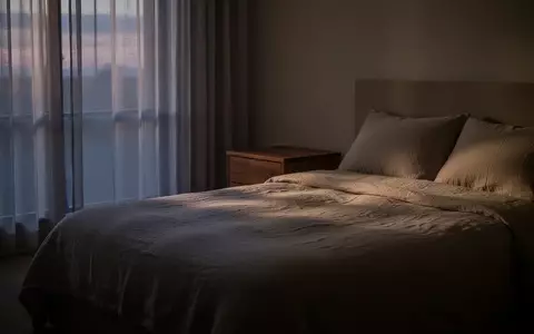 Ein minimalistisch eingerichtetes, ruhiges Schlafzimmer in der Abenddämmerung mit weichem Licht und einer ordentlichen Bettdecke. - Foto: über boerse-global.de