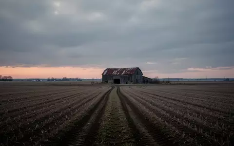 Ein einsamer Feldweg führt in der Dämmerung auf eine verwitterte Scheune in einer weitläufigen Agrarlandschaft zu. - Foto: über boerse-global.de
