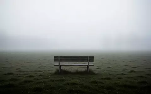Eine einsame, leere Parkbank auf einer nebligen Wiese in einer ruhigen, melancholischen Landschaft. - Foto: über boerse-global.de