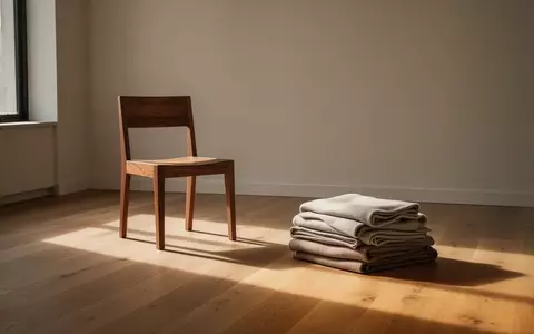Ein minimalistisch eingerichteter Raum mit einem leeren Stuhl und sauber gefalteter Kleidung auf einem Holzboden. - Foto: über boerse-global.de