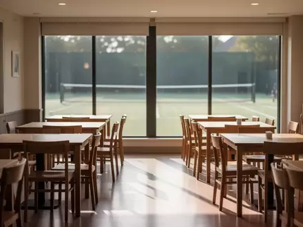 Ein leerer Gastraum in einem Vereinsheim mit Holztischen und Stühlen vor großen Fensterfronten mit Blick auf eine Tennisanlage. - Foto: über boerse-global.de