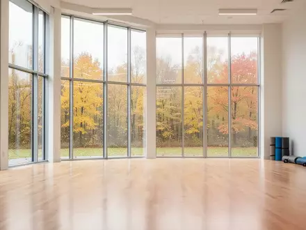 Ein heller, leerer Gymnastikraum mit Holzboden und Blick auf herbstliche Bäume durch große Fensterfronten. - Foto: über boerse-global.de