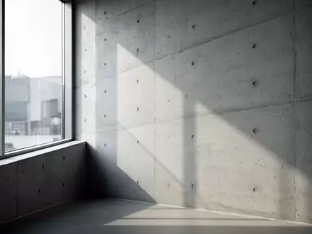 Ein minimalistischer Innenraum mit klaren architektonischen Linien und weichem Tageslicht, das geometrische Schatten auf eine Betonwand wirft. - Foto: über boerse-global.de