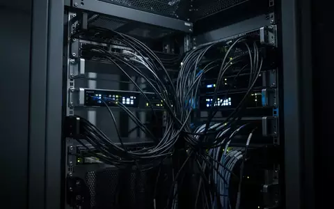 Dunkler Serverraum mit leuchtenden Statusanzeigen und einem Gewirr aus Glasfaserkabeln. - Foto: über boerse-global.de
